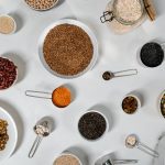 Flat lay of various dried grains, seeds, and beans arranged in bowls and measuring cups on marble.